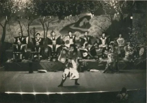 A black and white photo of a traditional dance performance with musicians in the background, possibly from a cultural or folk event.