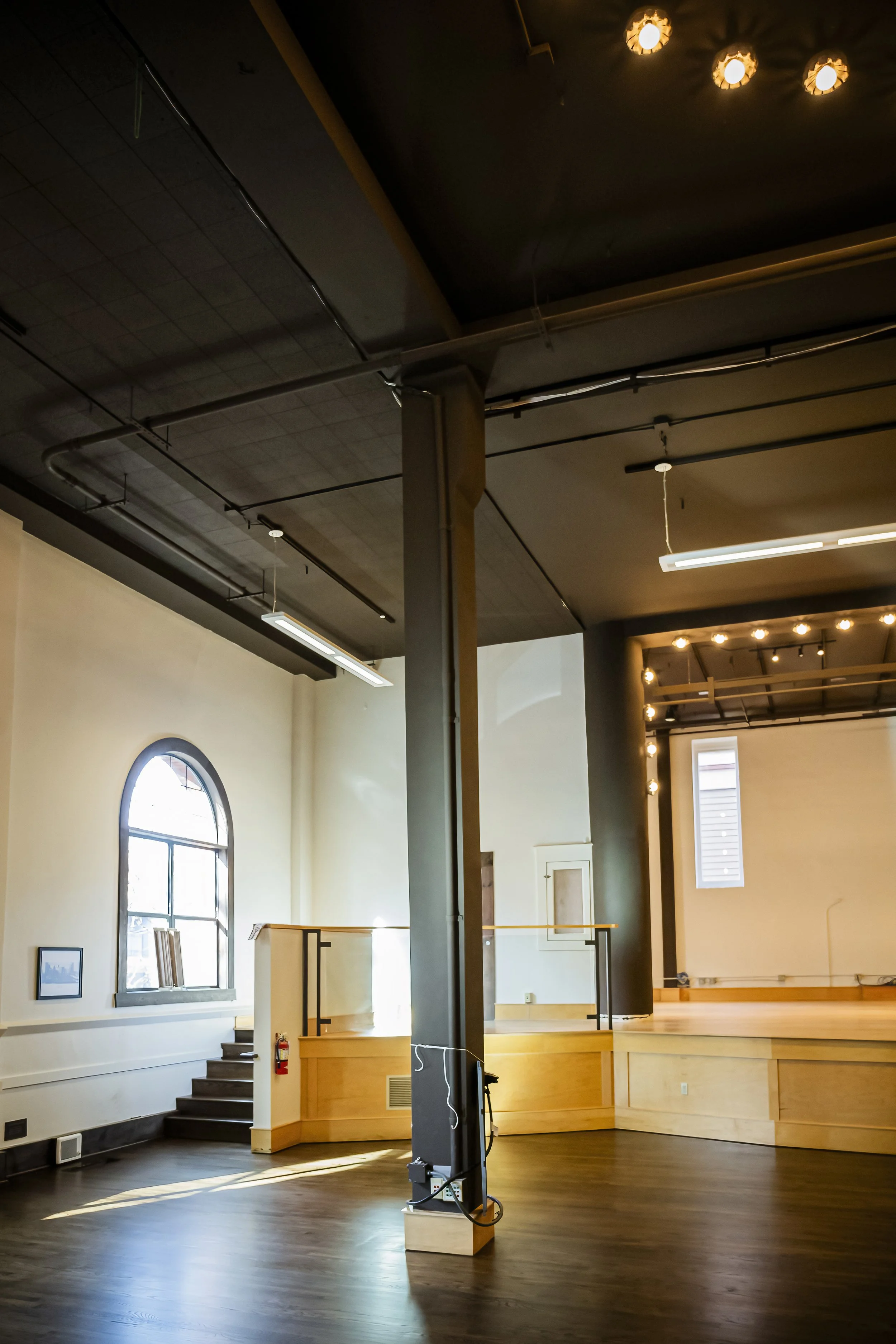 Empty interior space with a stage, large arched window, wooden floors, and ceiling lights, suggesting a performance or event venue.