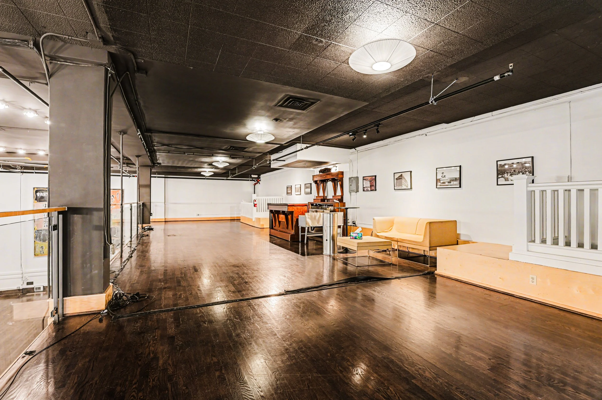 An empty, spacious indoor room with dark hardwood flooring, white walls decorated with framed photos, and a black ceiling with round light fixtures. On the right side, there's a yellow sofa, a glass coffee table with a box of tissues, and a wooden re