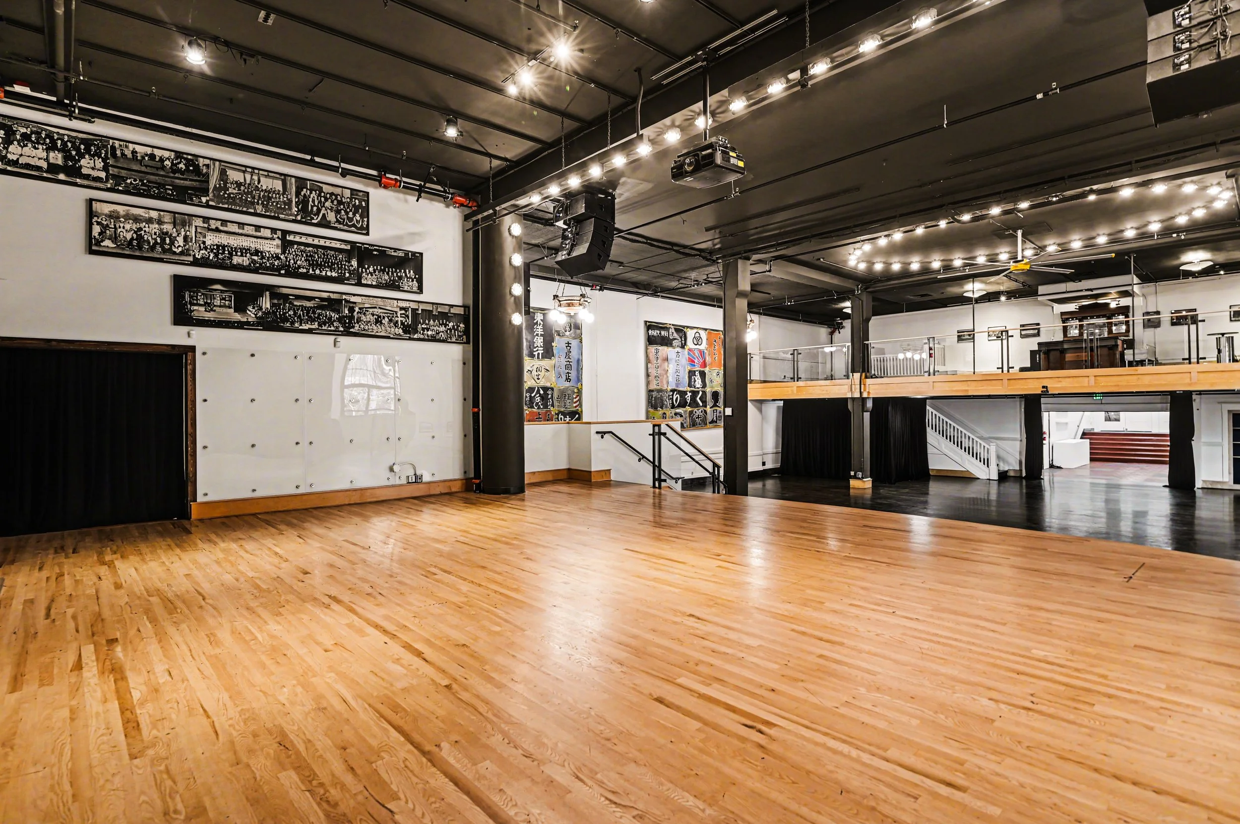 Nippon kan theatre, seattle, washington,Empty dance or event hall with wooden and dark flooring, black ceiling with lights, photo of black and white group pictures on the wall, stage area with colorful posters, staircases, and a second-floor balcony.