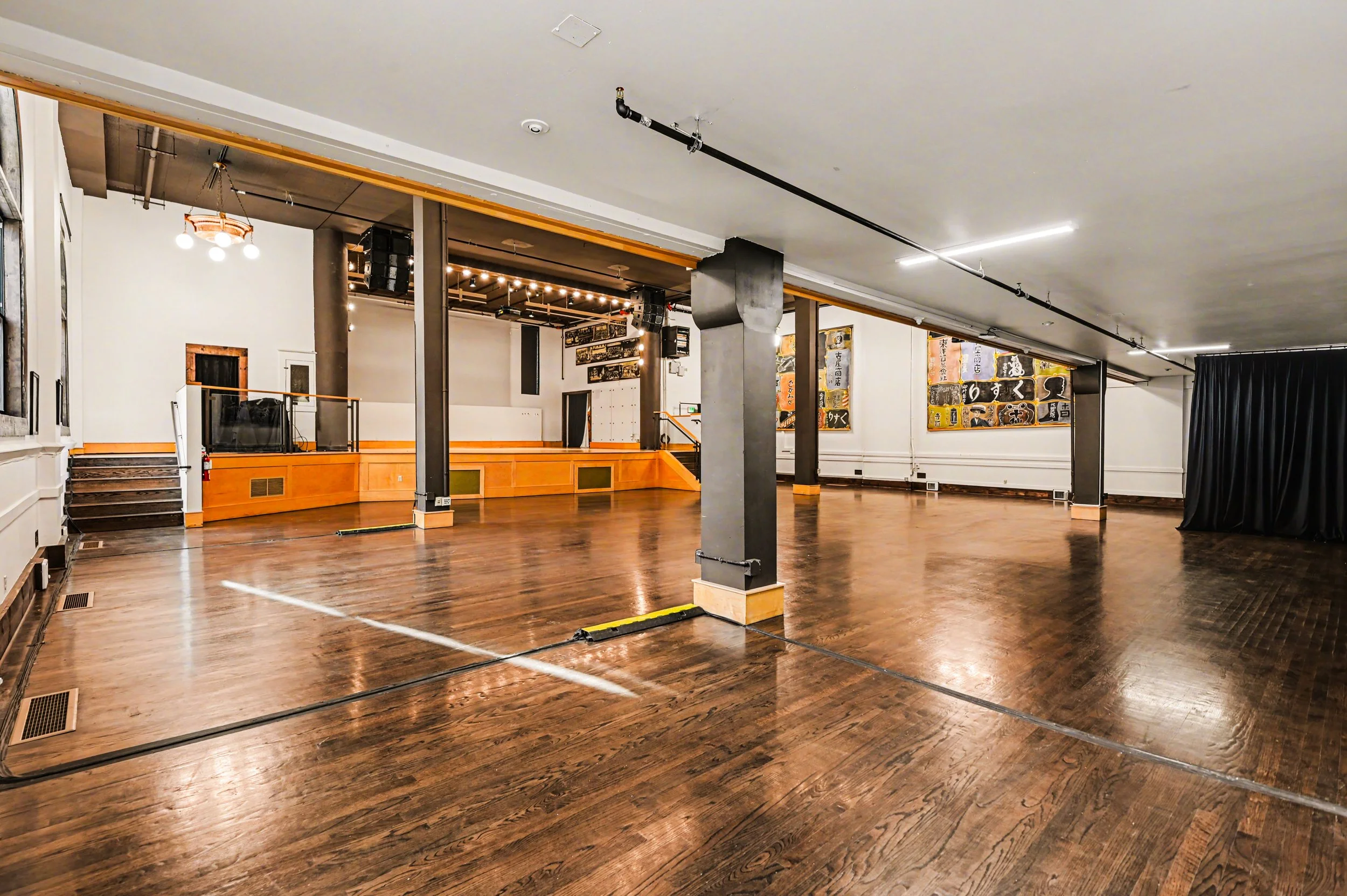 Nippon kan theatre, seattle, washington, event space, wedding,Empty dance or event hall with wooden floor, small stage area at the back, and black curtains on the right side. Walls decorated with artwork, and ceiling with exposed piping and lighting.