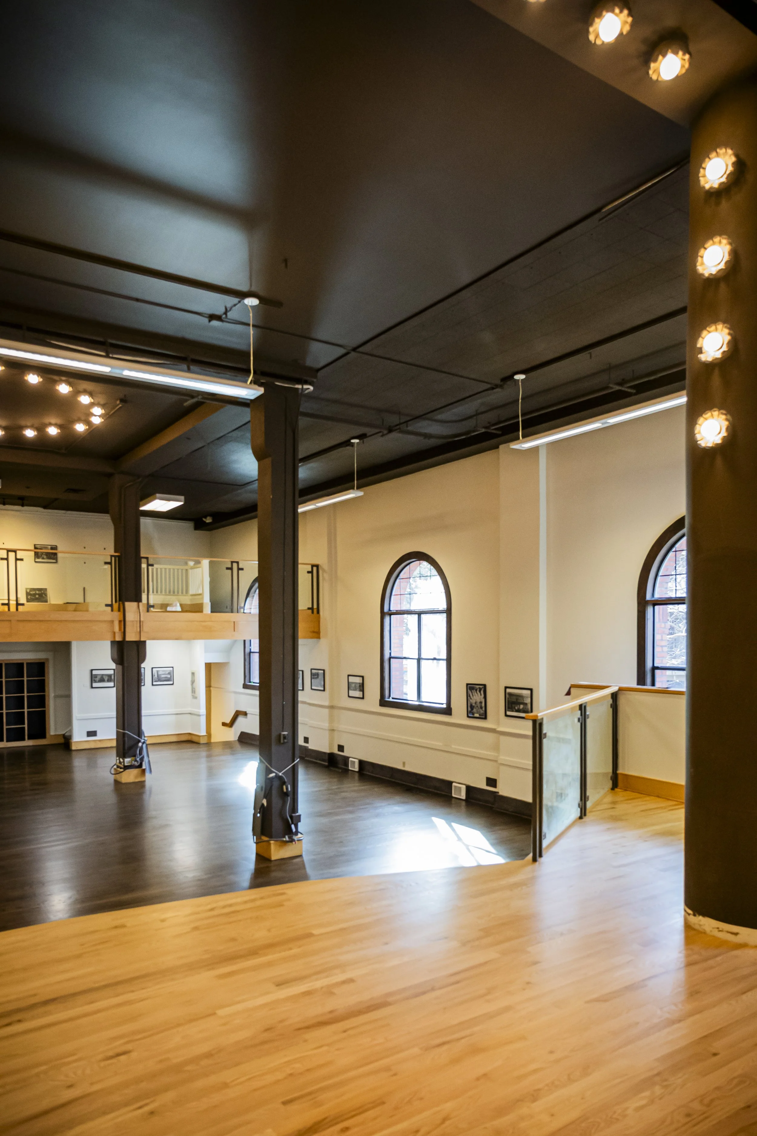 Empty multi-level studio or gallery space with wooden floors, white walls, arched windows, and framed pictures. Elevated balcony with glass railing and overhead lighting.