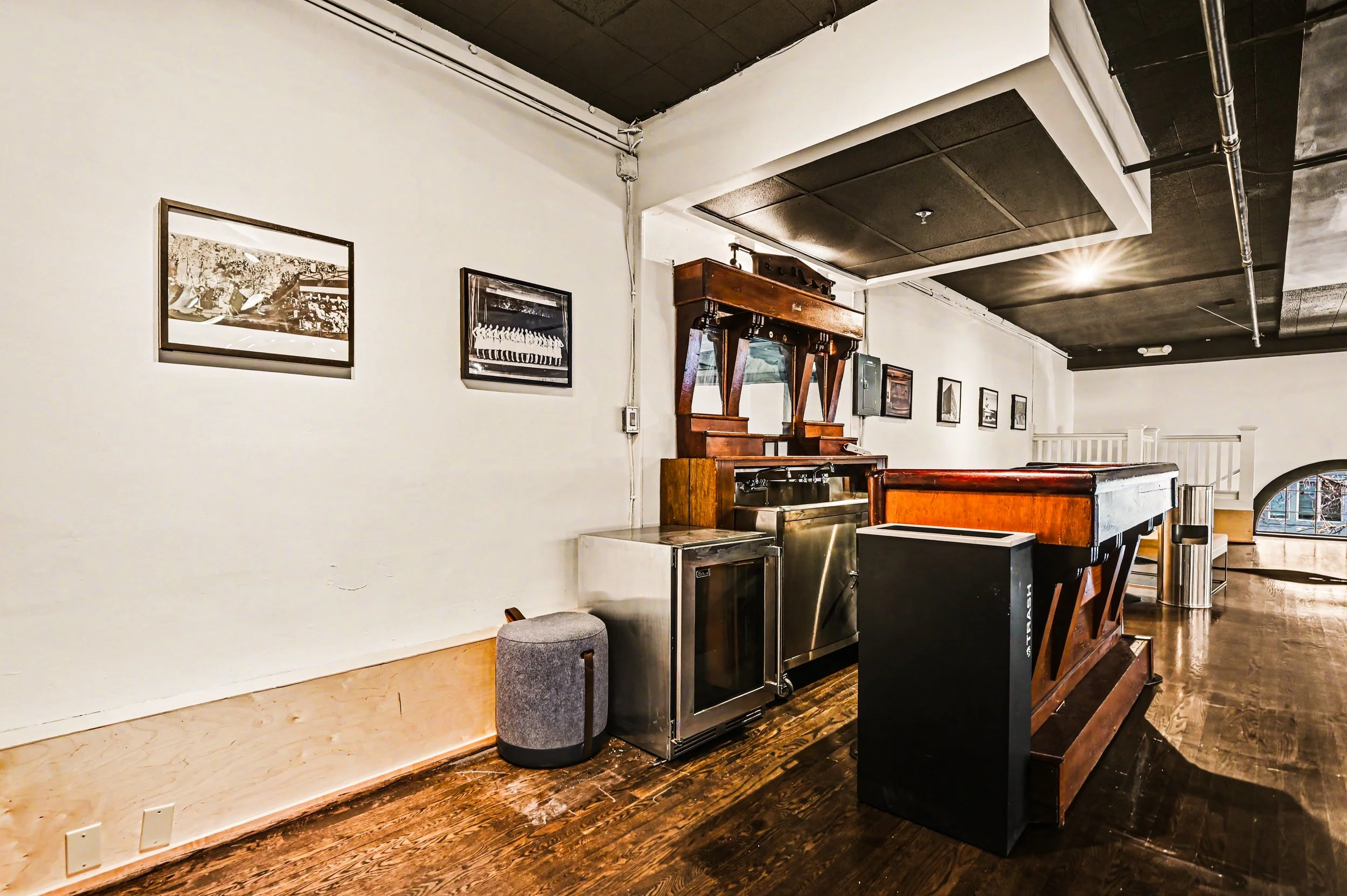 Interior of a cafe or restaurant with wooden flooring, white walls, and black ceiling panels. The wall is decorated with framed black-and-white photographs. There is a vintage wooden sideboard, stainless steel appliances, and a wooden counter. A stoo