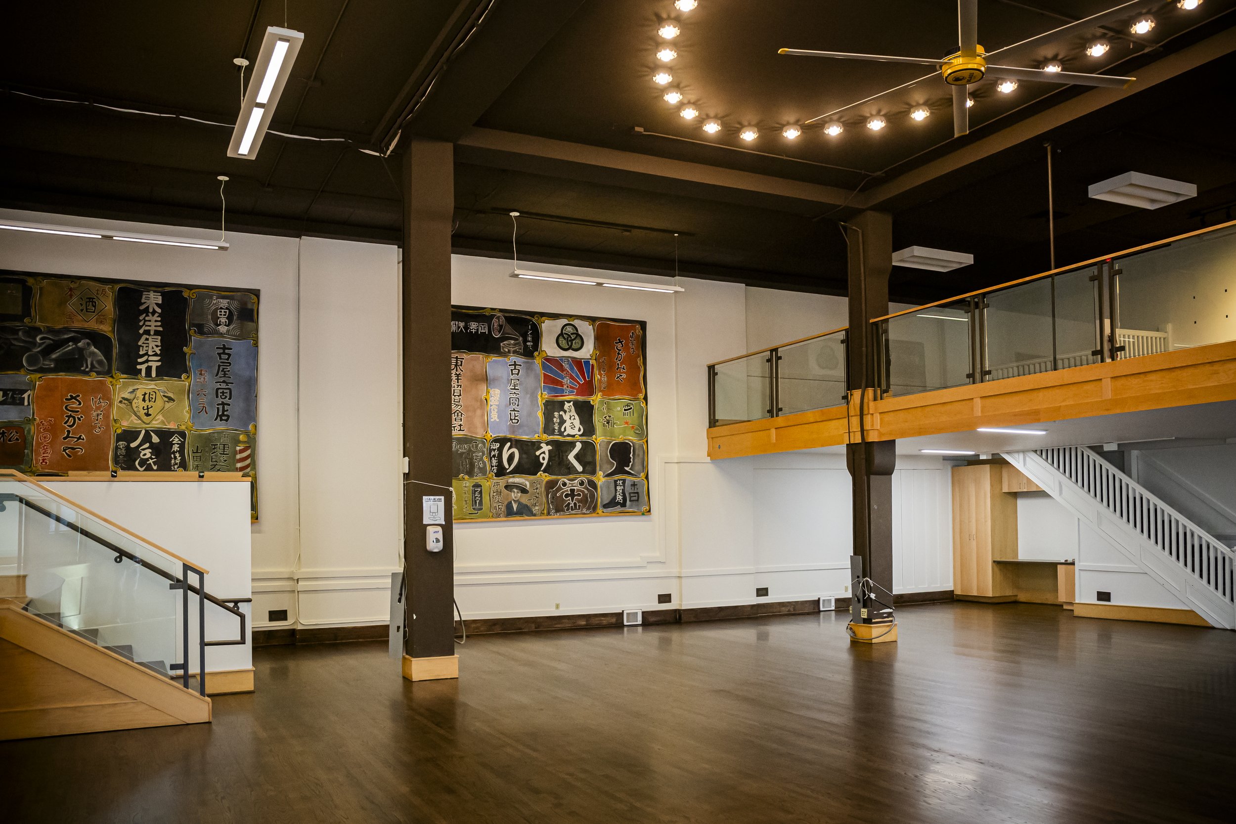 Empty interior space with dark wood flooring, white walls, and wooden accents, including a staircase and a balcony, with Japanese posters on the wall and ceiling lights.