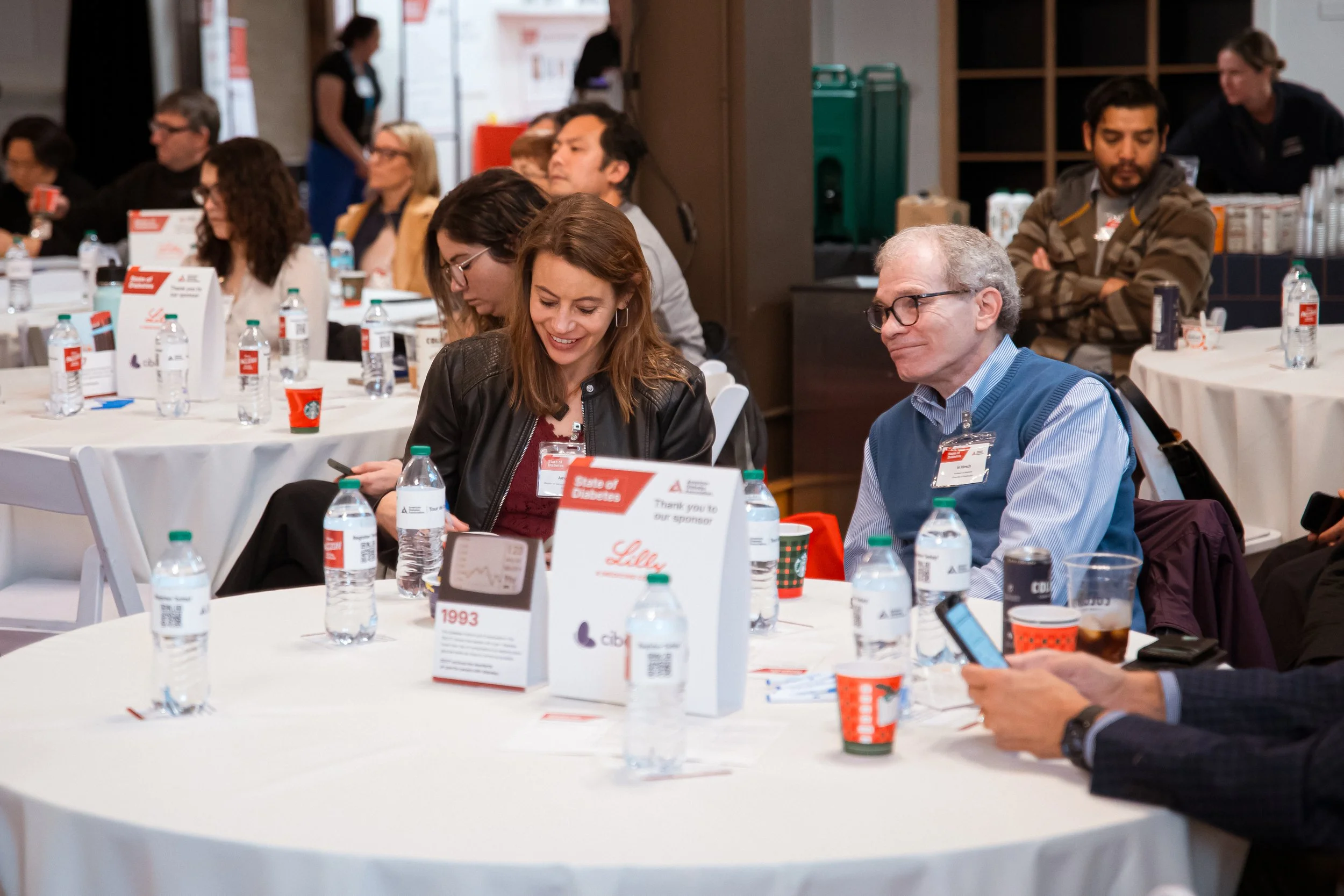 People sitting at round tables with water bottles, cups, and event materials, attending a conference or seminar in a well-lit room.