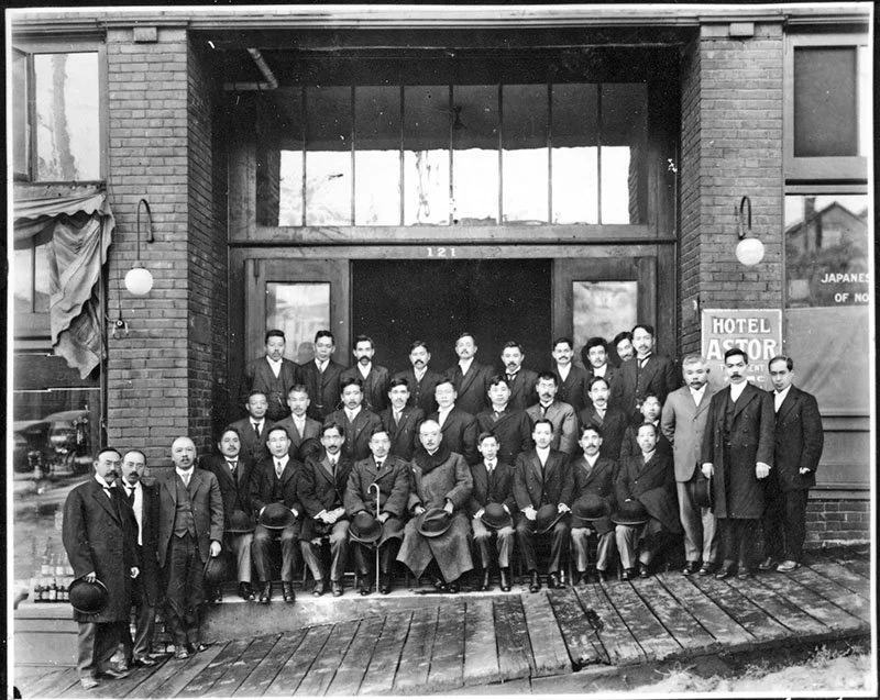 Black and white photo of a group of men in suits posing outside a building labeled HOTEL ASTOR.