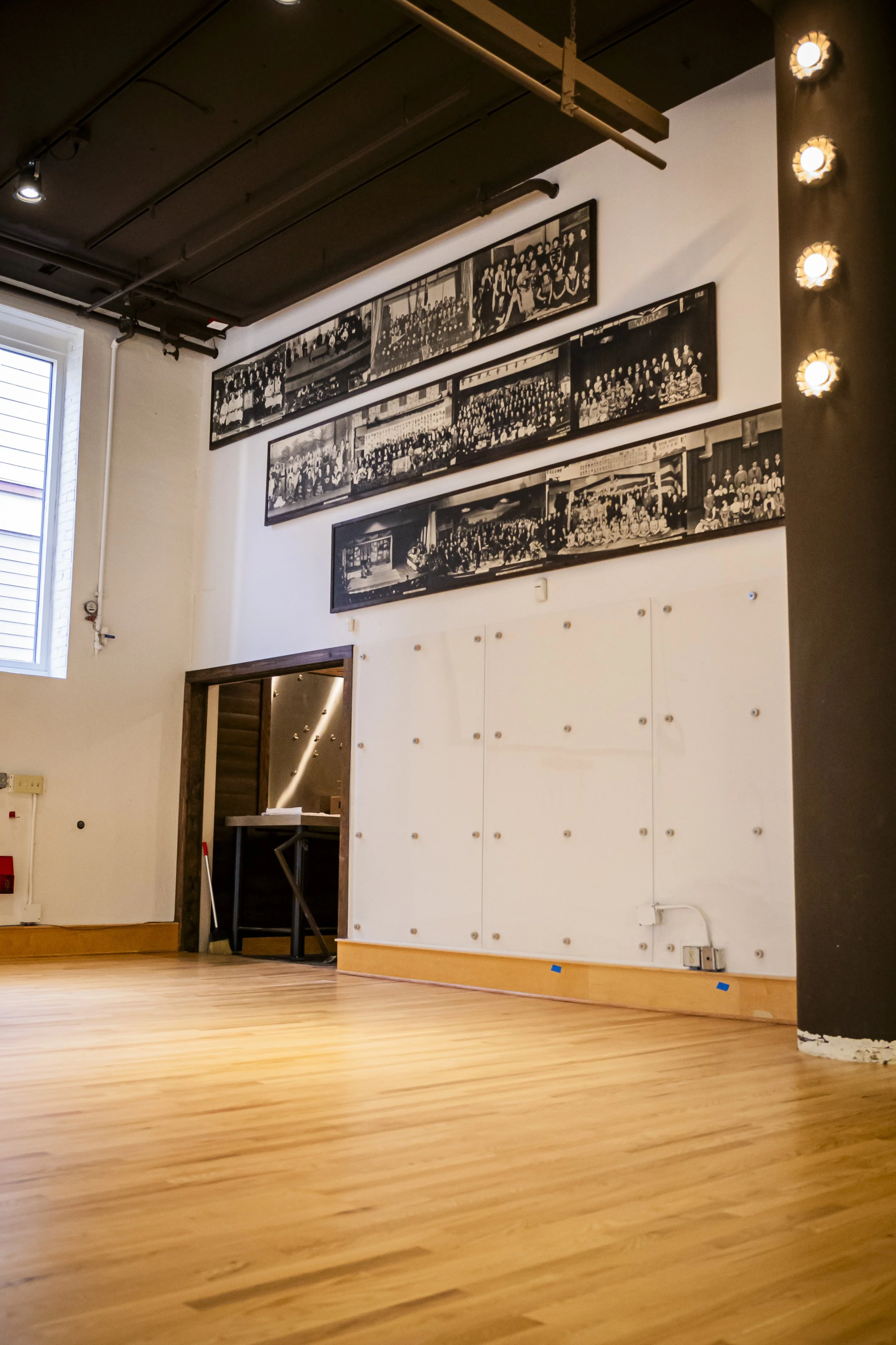 Empty room with hardwood floor, white wall with large black-and-white group photos, black ceiling, and exposed pipes. There is a window on the left with blinds and a doorway leading to another space.
