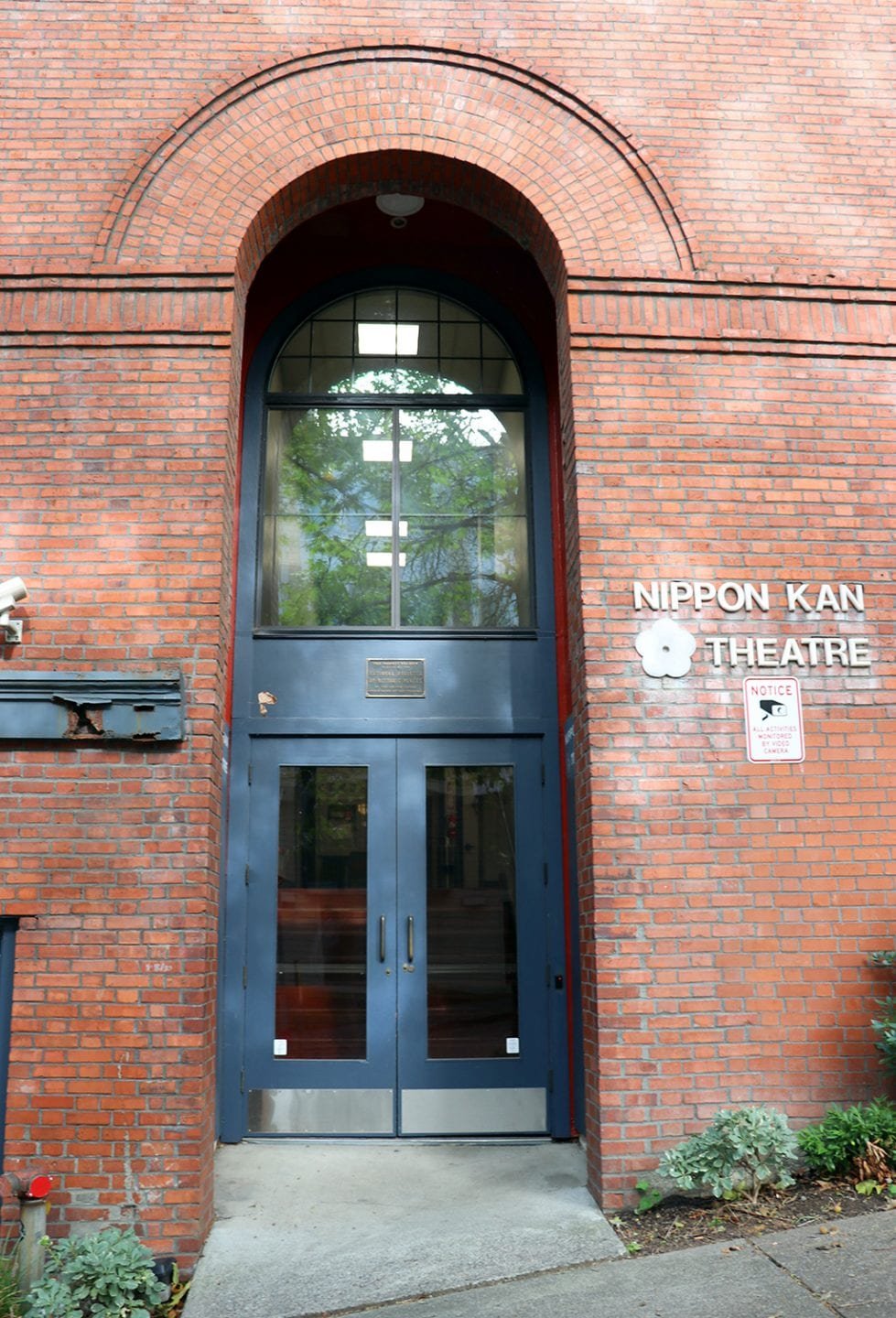 Brick building entrance with glass double doors, sign reading 'Nippon Kan Theatre,' and a notice sign with security camera icon.