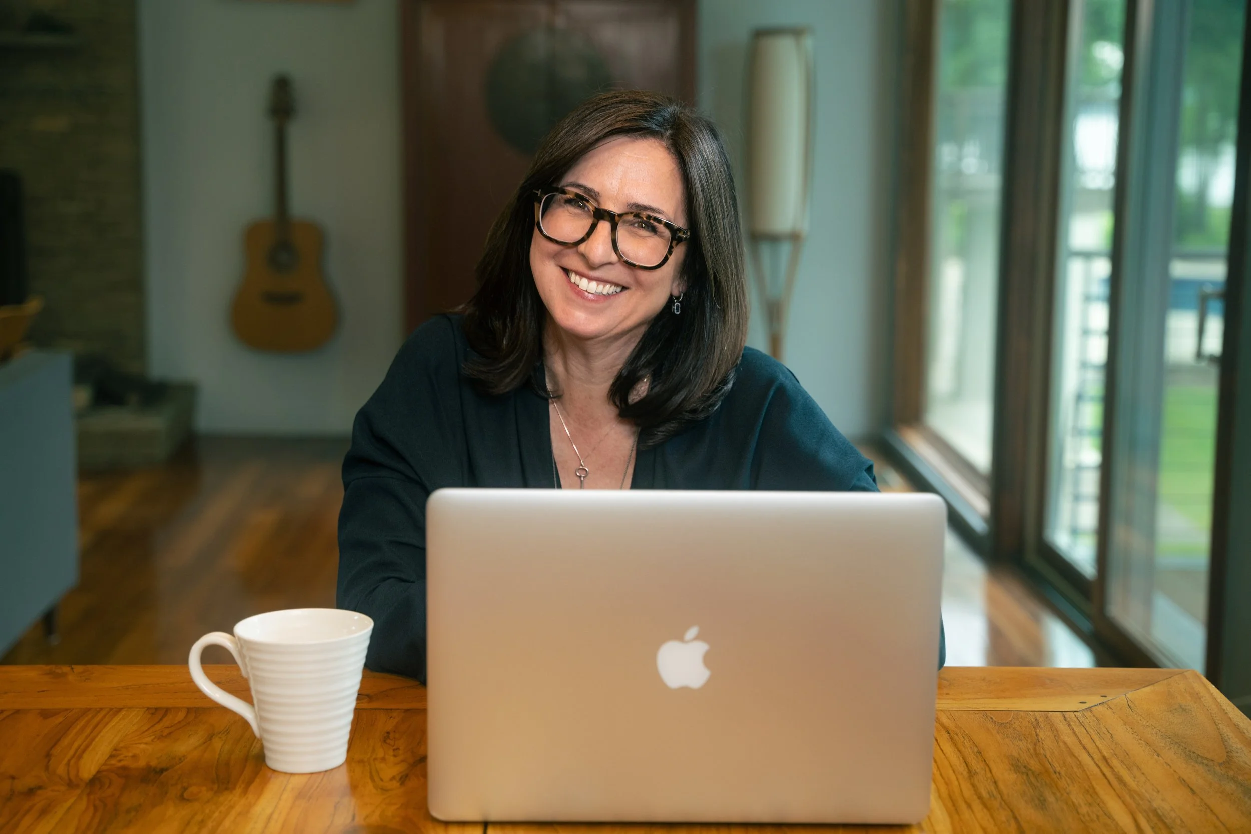 A woman with shoulder-length brown hair, wearing glasses and a black top, sitting at a wooden table with an open MacBook and a white mug, smiling at the camera in a bright room with large windows and a guitar in the background.