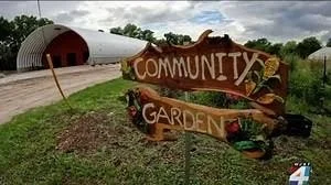 A wooden sign reads 'Community Garden' with a garden bed in front and a tunnel structure in the background under a cloudy sky.