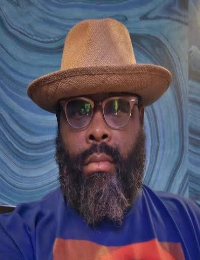 A man with a beard wearing sunglasses, a brown hat, and a blue shirt, against a swirled blue patterned background.