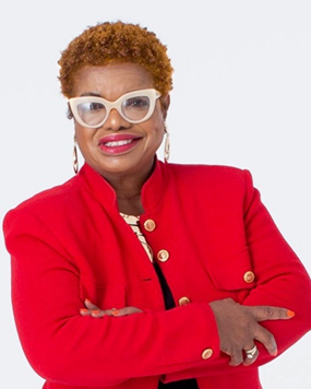 A confident woman with short curly red hair wearing white glasses and a red blazer, smiling with arms crossed against a white background.