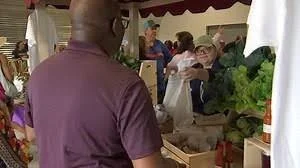 Person shopping at a farmers market with various fresh produce and other shoppers in the background.