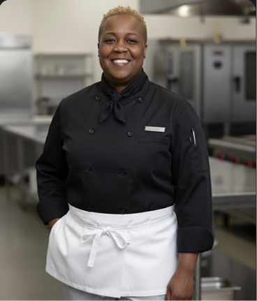 Smiling female chef in black chef's jacket and white apron standing in commercial kitchen.