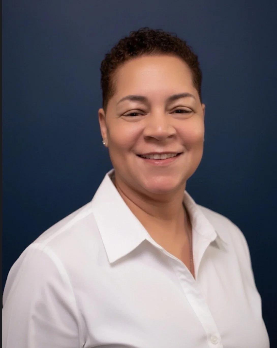 A woman with short curly hair, smiling, wearing a white shirt, against a dark blue background.