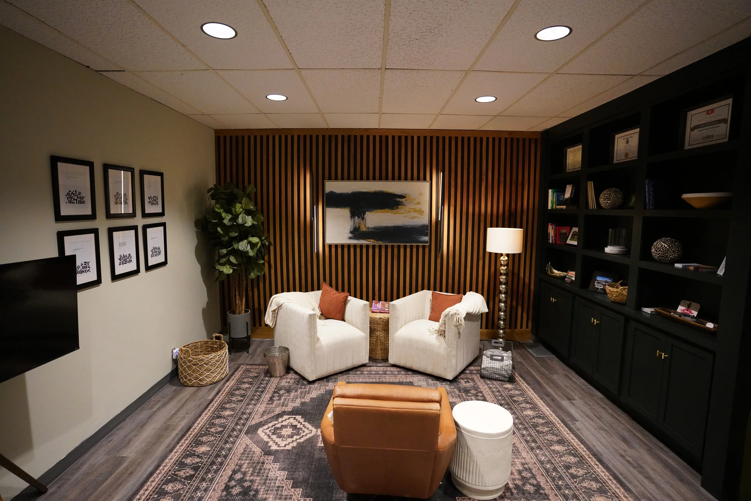 Cozy office or lounge room with two white armchairs, a brown leather chair, an abstract wall art, a tall floor lamp, a large black bookshelf with decor items, framed pictures, and a patterned rug.