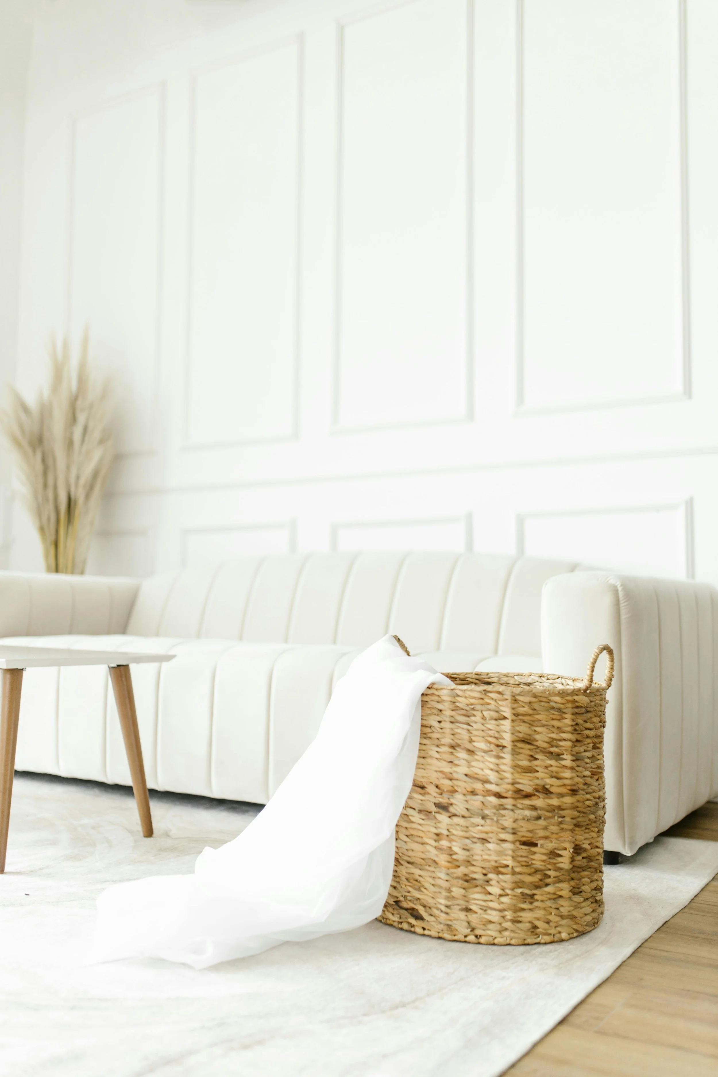 A white sofa with black legs, a small wooden table, a wicker basket with a white cloth, and a beige rug in a bright living room with white paneled walls and dried pampas grass in a vase.