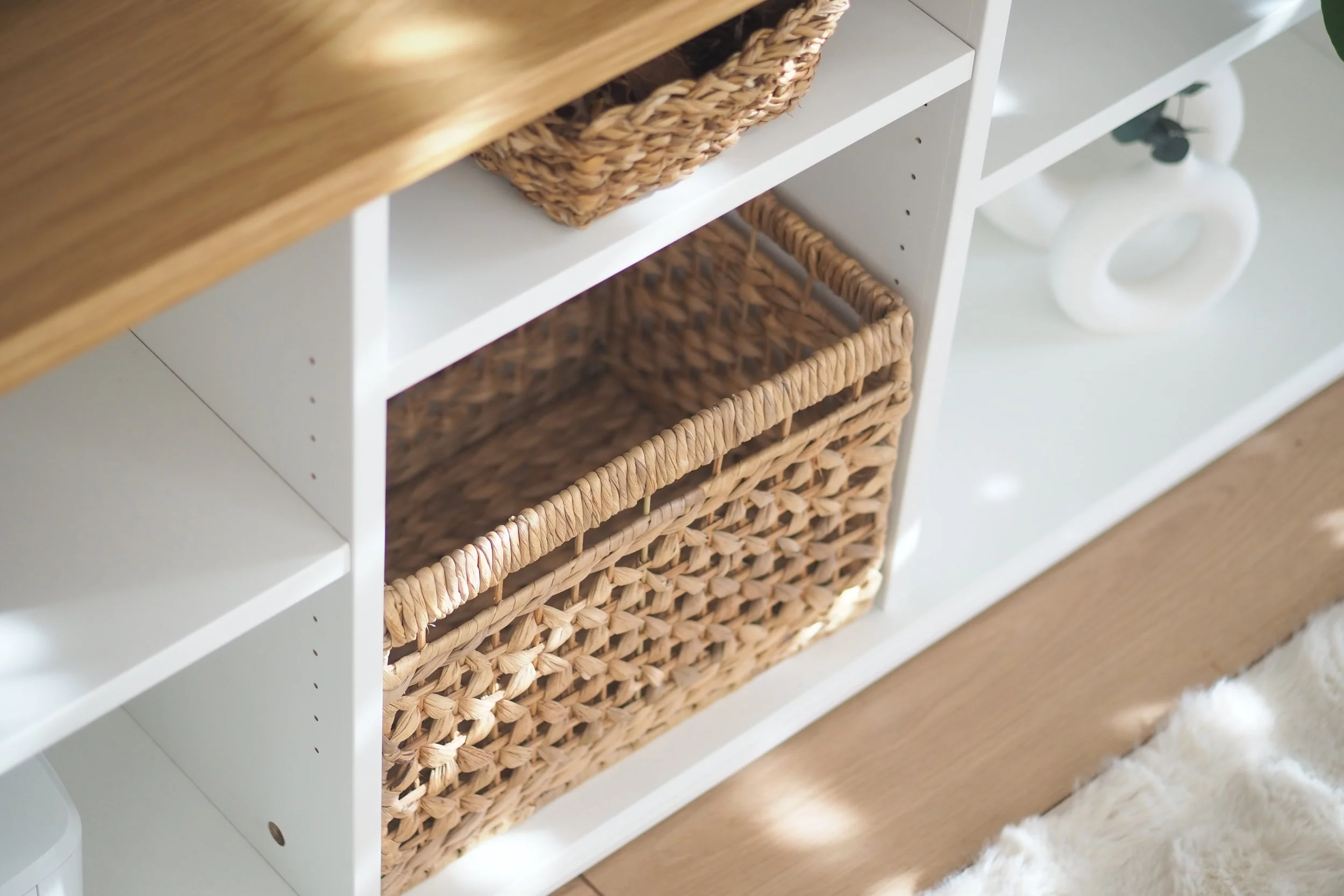 A white shelving unit with woven baskets on the shelves, situated on a wood floor with a white fluffy rug nearby.