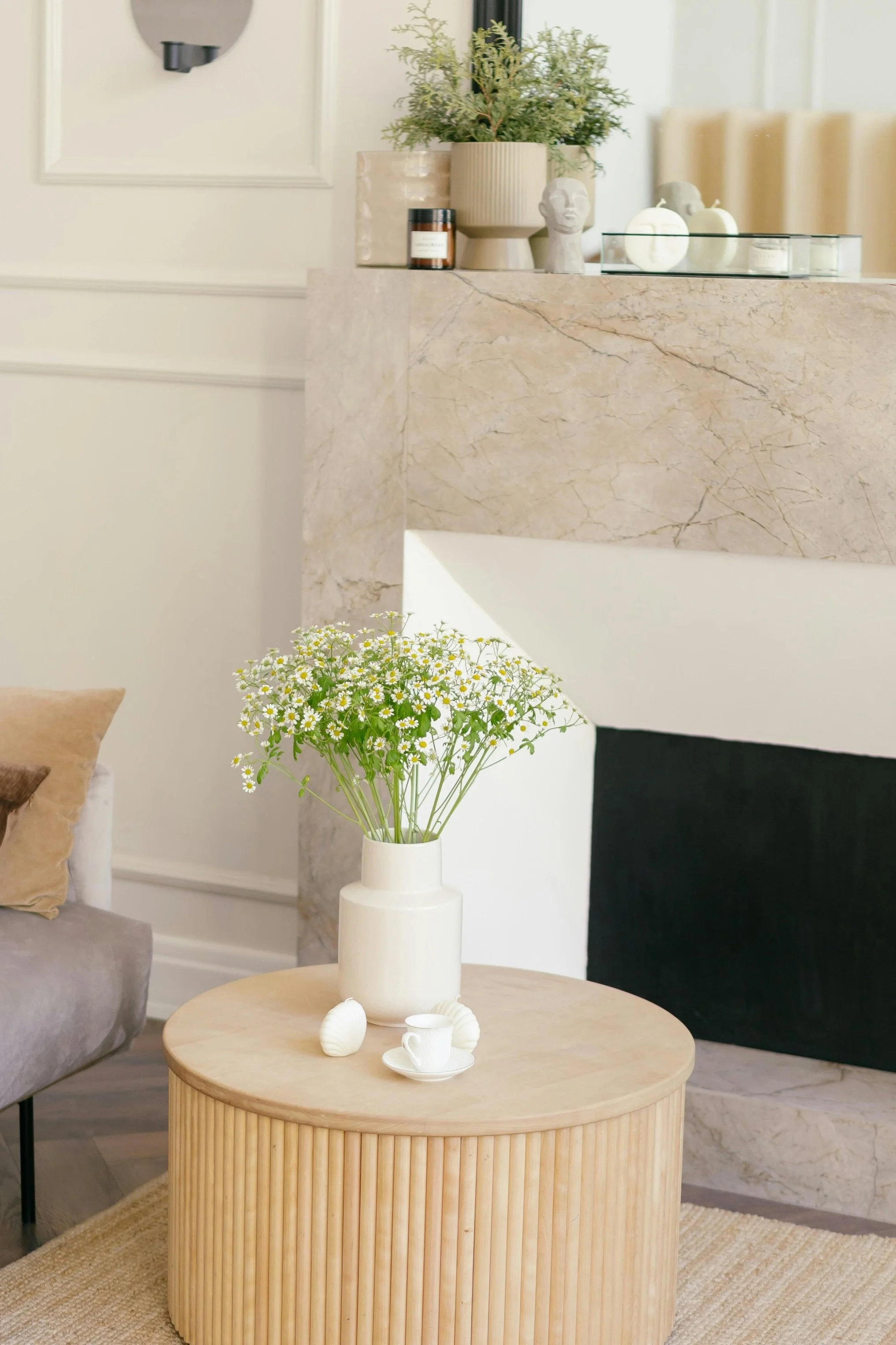 A round, ribbed wooden coffee table with a white vase holding a bouquet of daisies, a small white teacup, and shell-shaped decor. In the background, a beige sofa with a brown pillow, a marble fireplace mantle decorated with potted plants, head sculptures, and candles, and a wall with artwork and wall-mounted light fixtures.