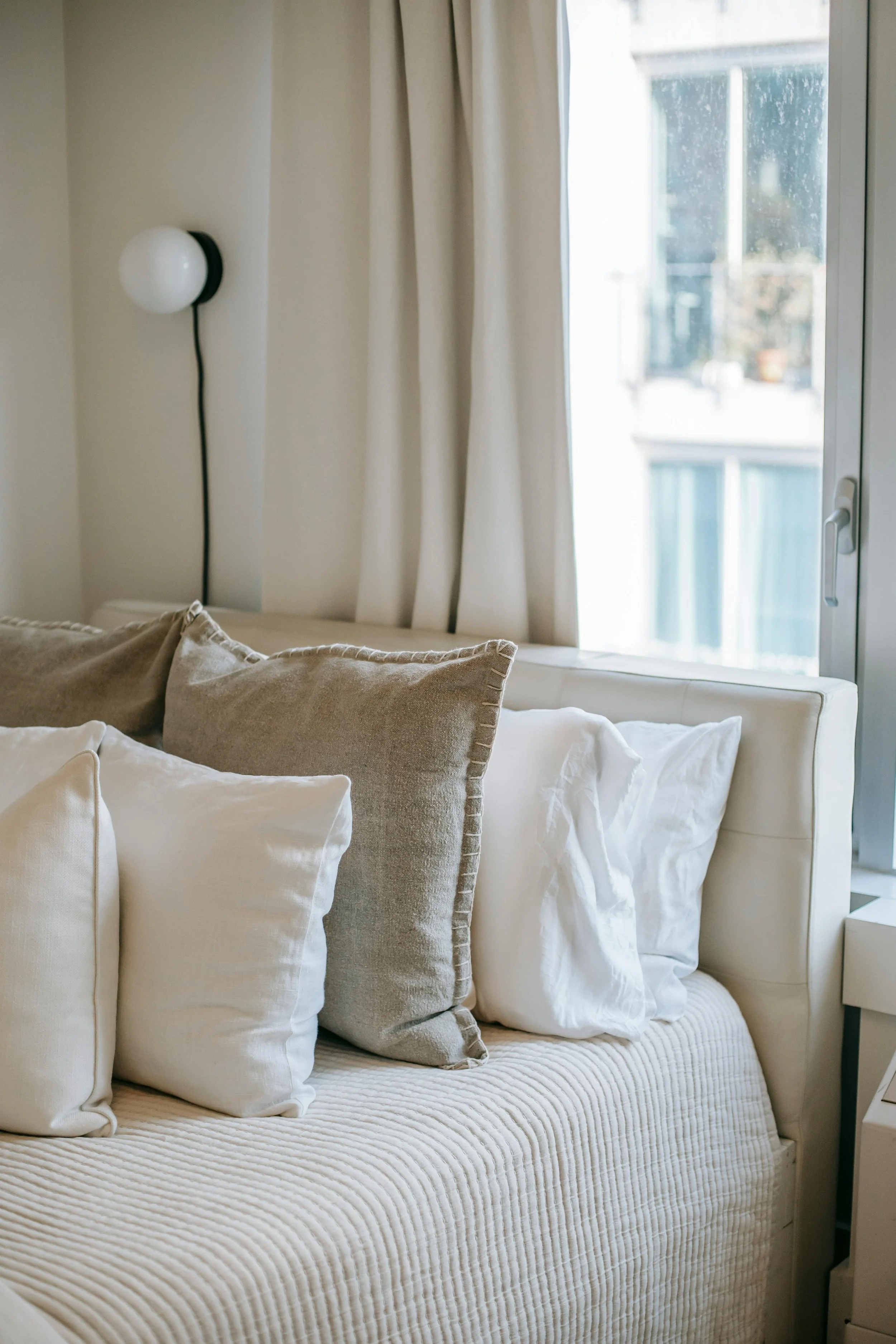 Bed with multiple beige and white pillows in front of a window with cream curtains, natural light coming through.