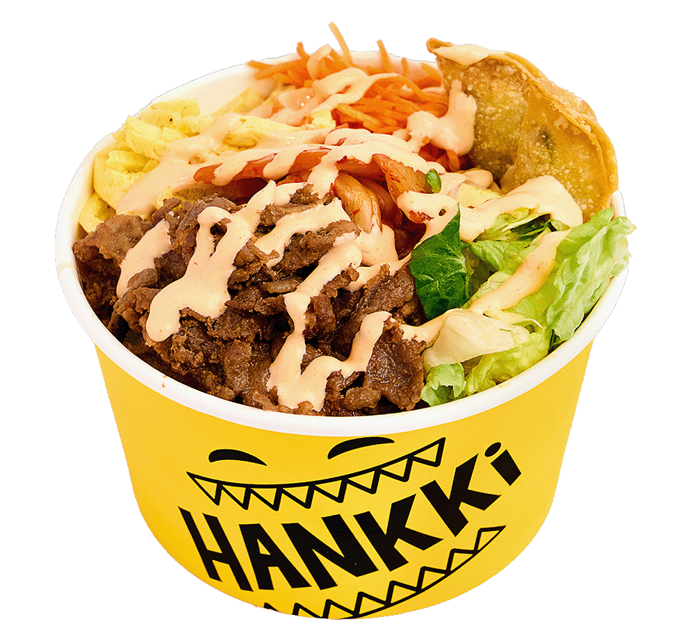 A yellow bowl with a happy face and the word 'HANKKI' contains a salad topped with grilled beef, shredded carrots, and drizzled with a creamy dressing.