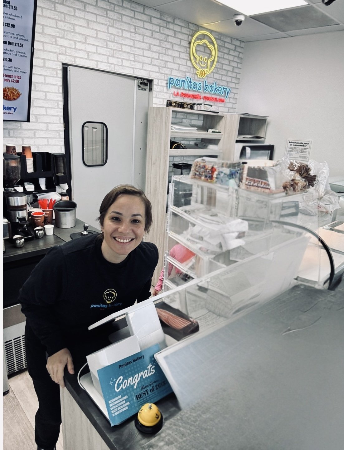 Smiling woman working at cafe counter inside Panitas Bakery, with bakery products, receipt box, and coffee-making area visible.