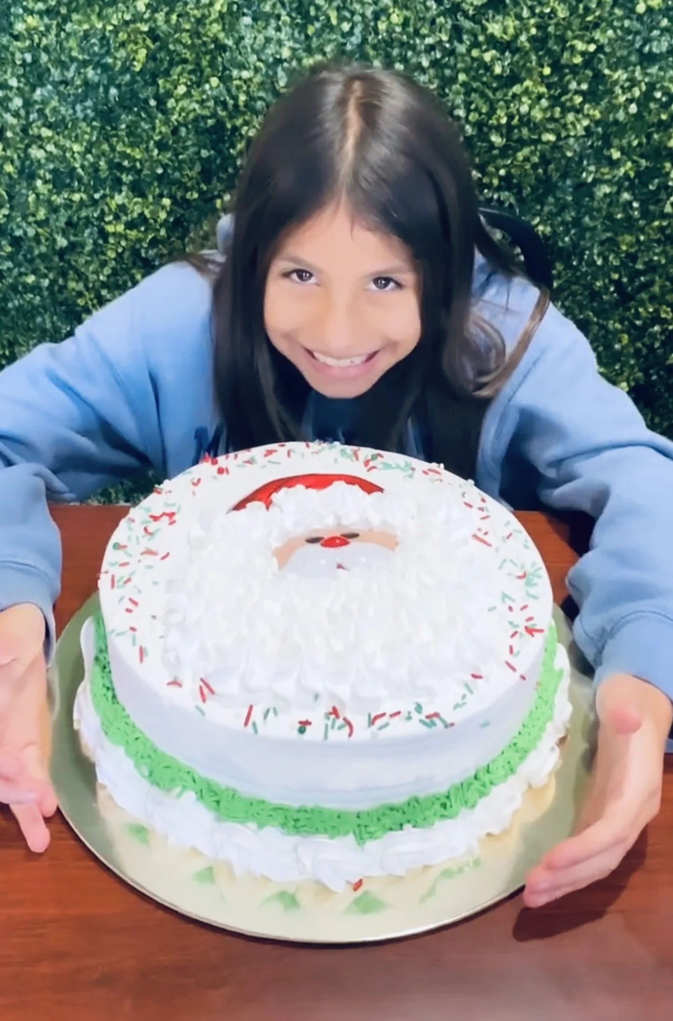 A young girl with long dark hair smiling, leaning over a Christmas cake with a Santa face decoration, on a wooden table in front of Christmas greenery.