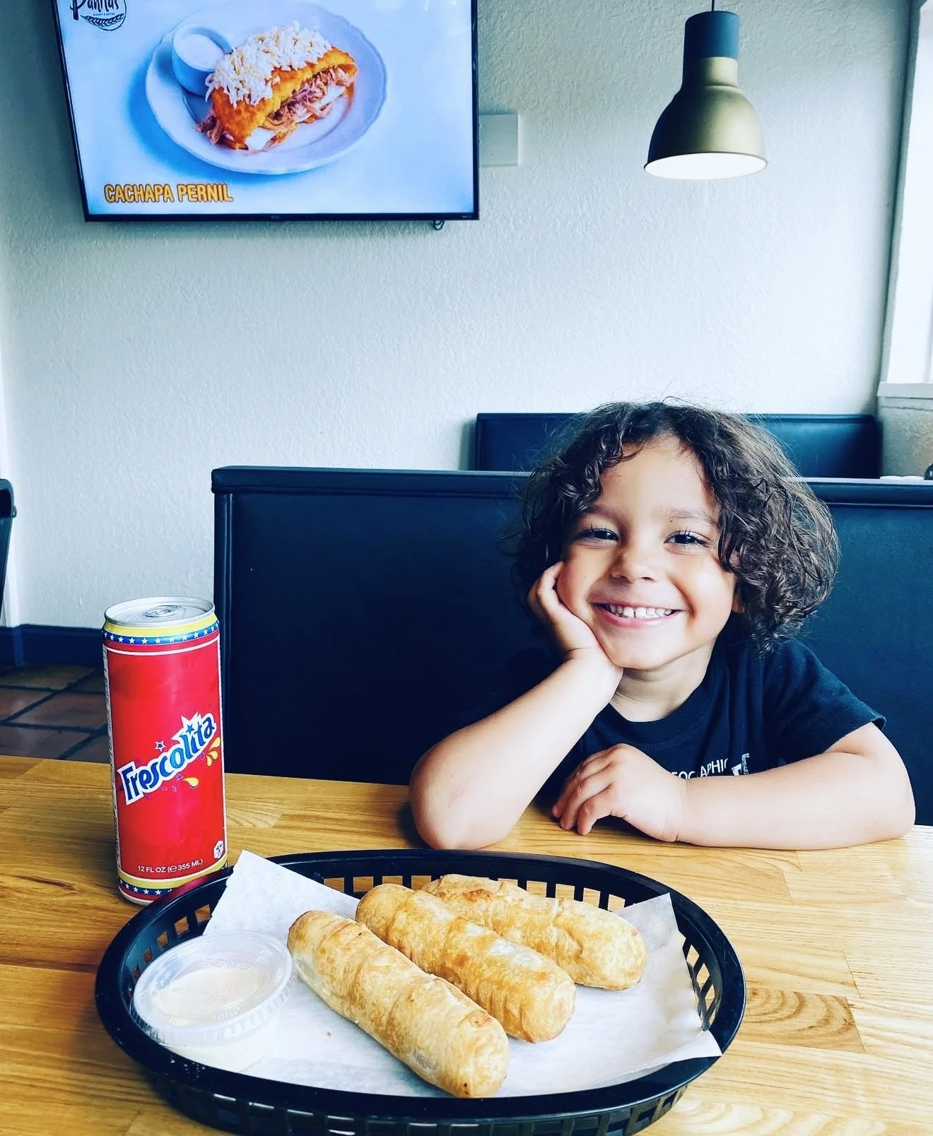 A young boy with curly hair smiling, sitting at a wooden table with a black basket of four fried taquitos, a small cup of dipping sauce, a can of Tecate soda, and a TV showing a Mexican dish called Cachapa Pernil in the background.