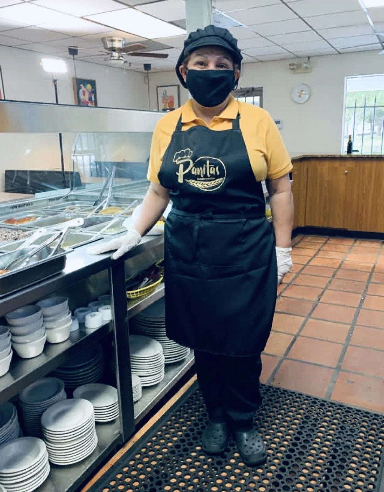 A woman in a yellow polo shirt and black apron with the word 'Pamis' on it, wearing a black face mask and gloves, standing in a restaurant or deli. She is near a counter with bowls and utensils, inside a restaurant with tiled flooring, ceiling fans, and some artwork on the walls.