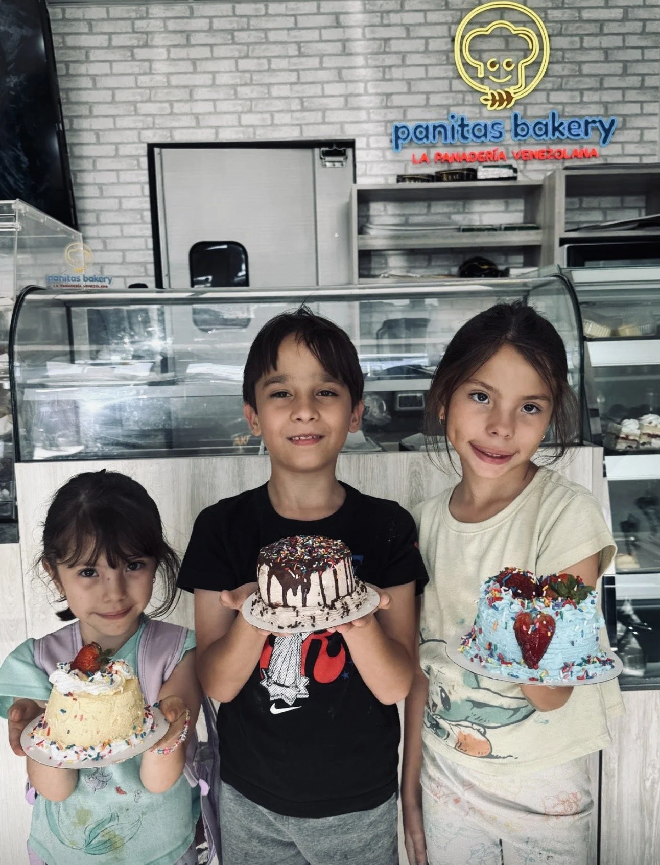Three children holding decorated cakes inside a bakery, with a sign reading 'panitas bakery' and a cartoon chef logo in the background.