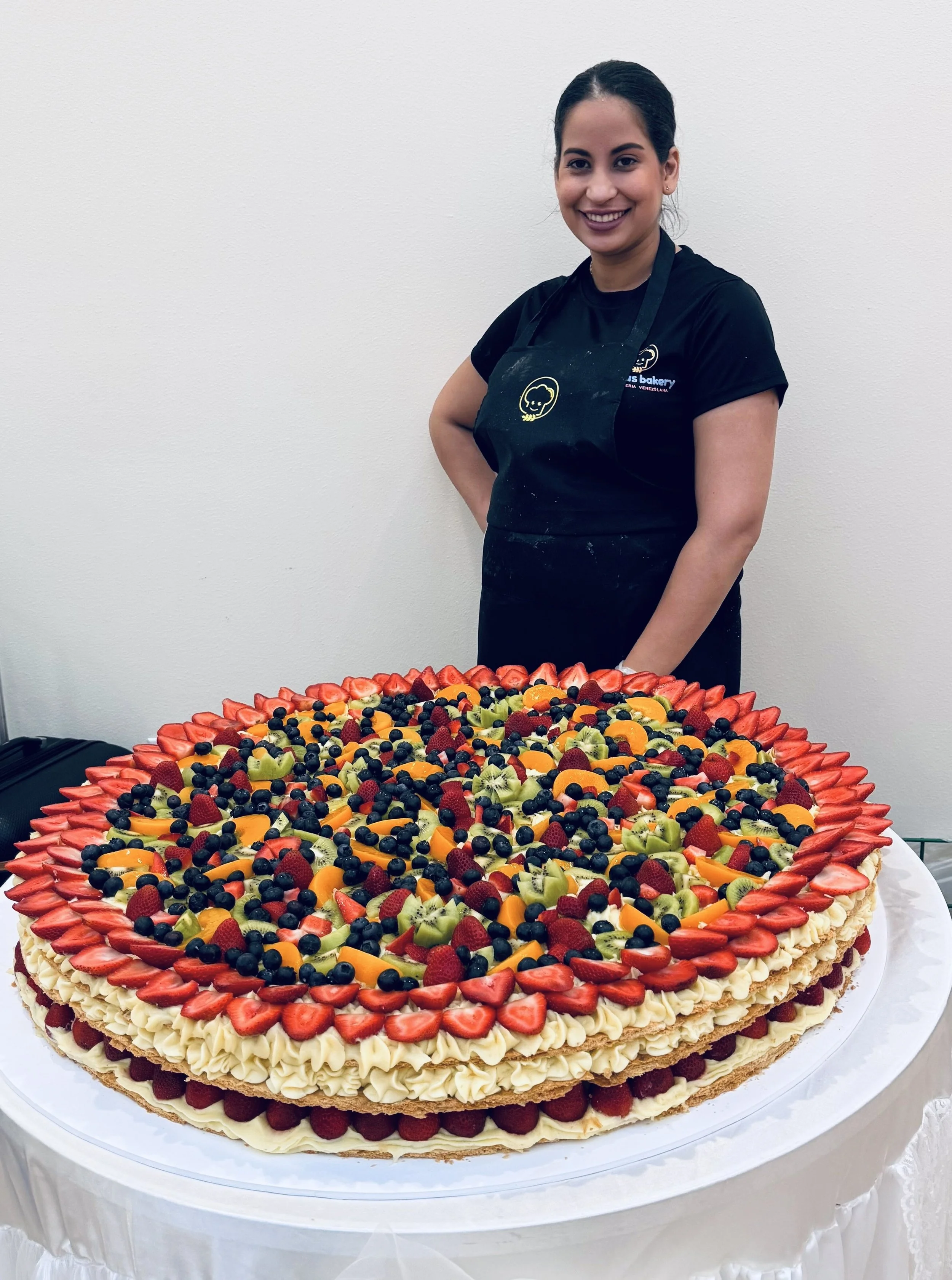 A woman with dark hair in a bun, wearing a black T-shirt and black apron, stands next to a large, round fruit-topped cake on a white table. The cake is decorated with strawberries, blueberries, kiwi, and other mixed fruits, with a cream border and sliced strawberries around the edge.