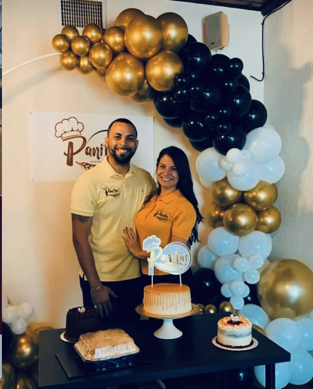 Celebrating a birthday with a man and woman standing behind a table with cakes, in front of a balloon arch in black, white, and gold.