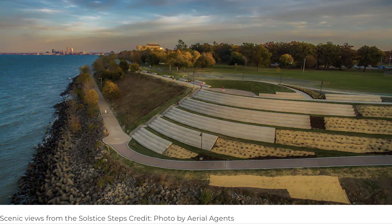 Aerial view of Solstice Steps along a lakeside park, with tiered steps leading to walking paths and green lawns, trees, and city skyline in the background at sunset.
