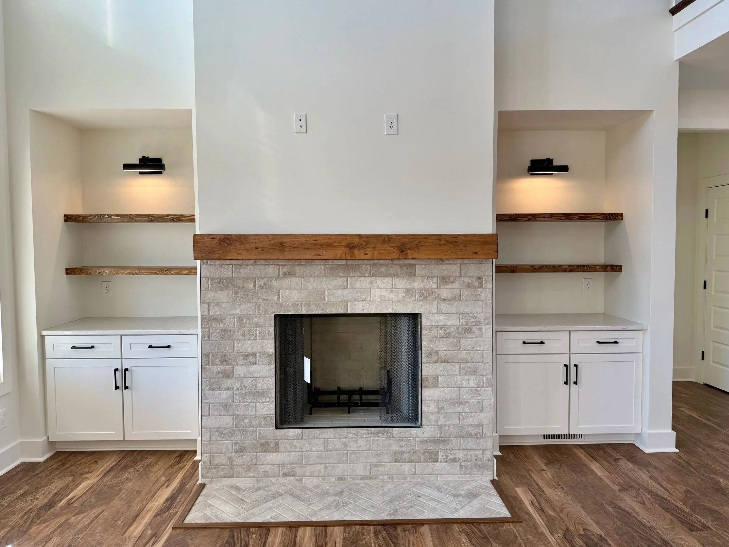 Living room fireplace with white brick surround, wooden mantel, built-in shelves with black wall sconces on each side, white cabinets below, and hardwood floors.