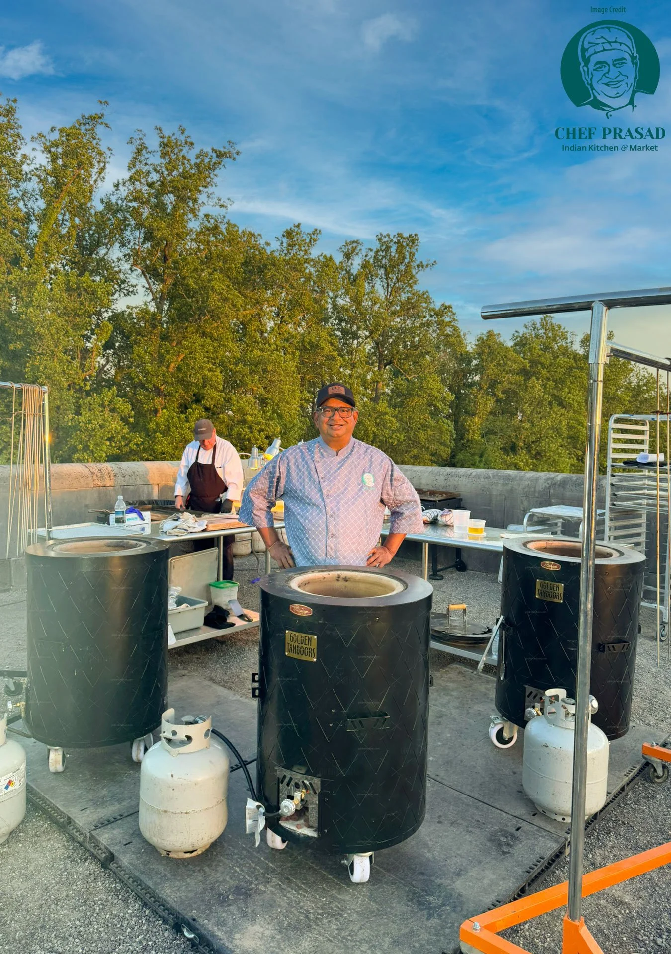 A chef standing in front of three large tandoors on a rooftop, with another person cooking in the background, trees and a blue sky visible.