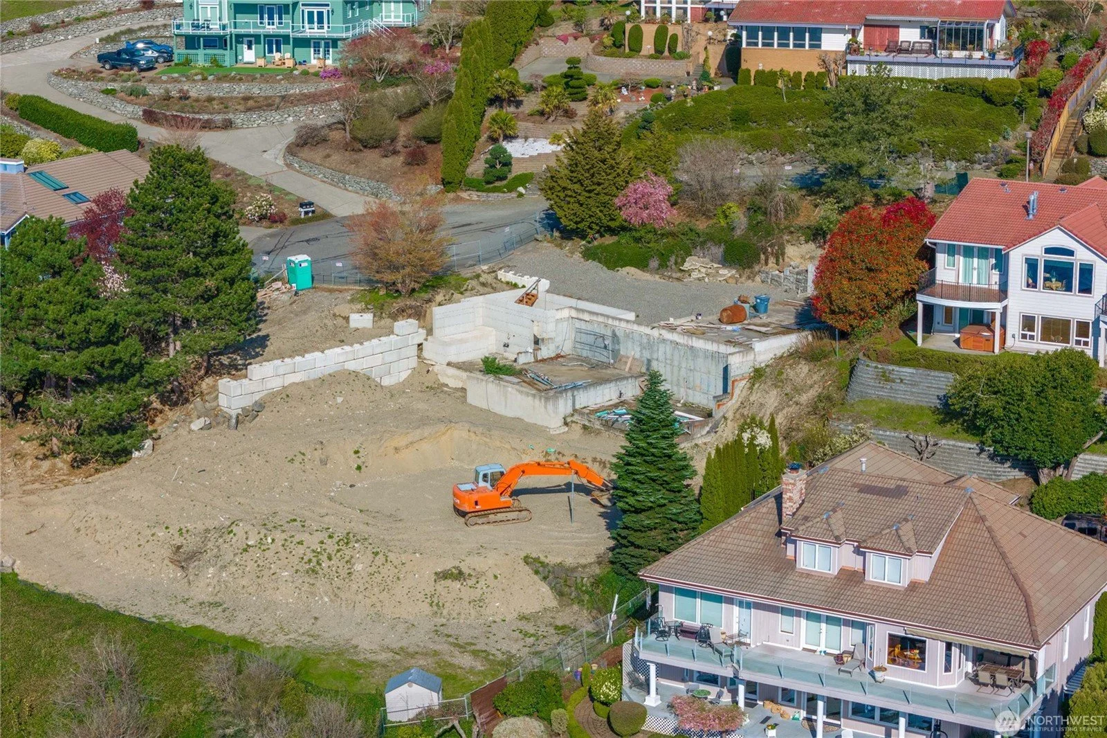 Aerial view of a residential neighborhood under construction with an excavator on a dirt lot, surrounded by houses with landscaped yards and trees.