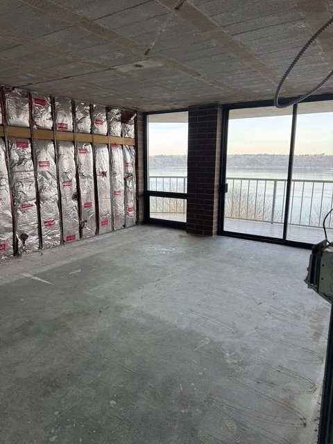 An unfinished room with insulation and brick wall, large windows showing a view of a body of water, and exposed ceiling and floor.