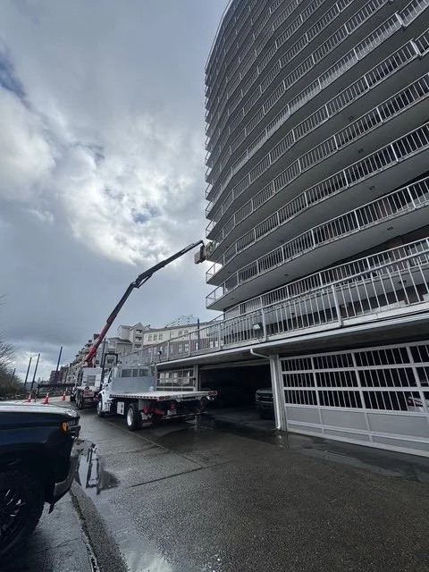 A construction vehicle with an extendable boom lift working on the exterior of a tall, curved residential building with multiple balconies, on a cloudy day with wet pavement.