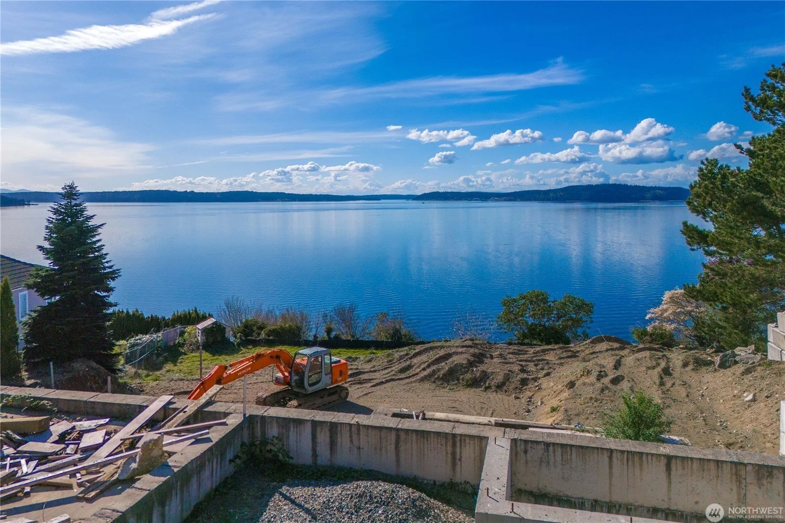 Construction site overlooking a large lake with a small excavator and dirt mounds, trees, and houses along the shoreline under a blue sky with clouds.