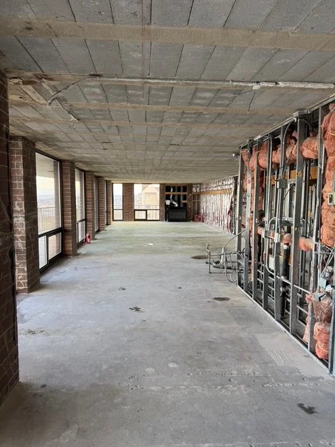Interior of a building under construction with exposed brick walls, insulation, and concrete floors, with large windows along one side.
