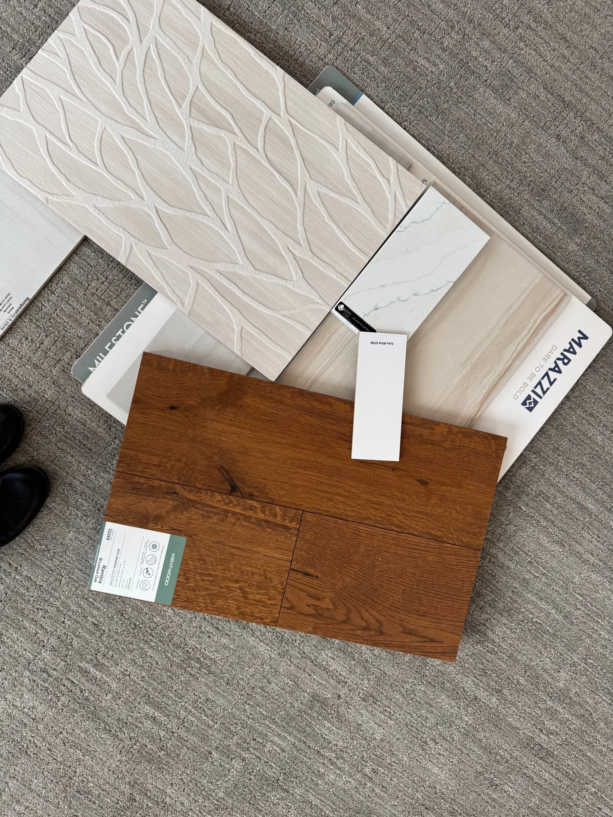 Samples of different types of flooring materials, including a wood-look tile, a white marble tile, a beige wood-look tile, and a textured white tile with a leaf pattern, displayed on the floor with some labels.