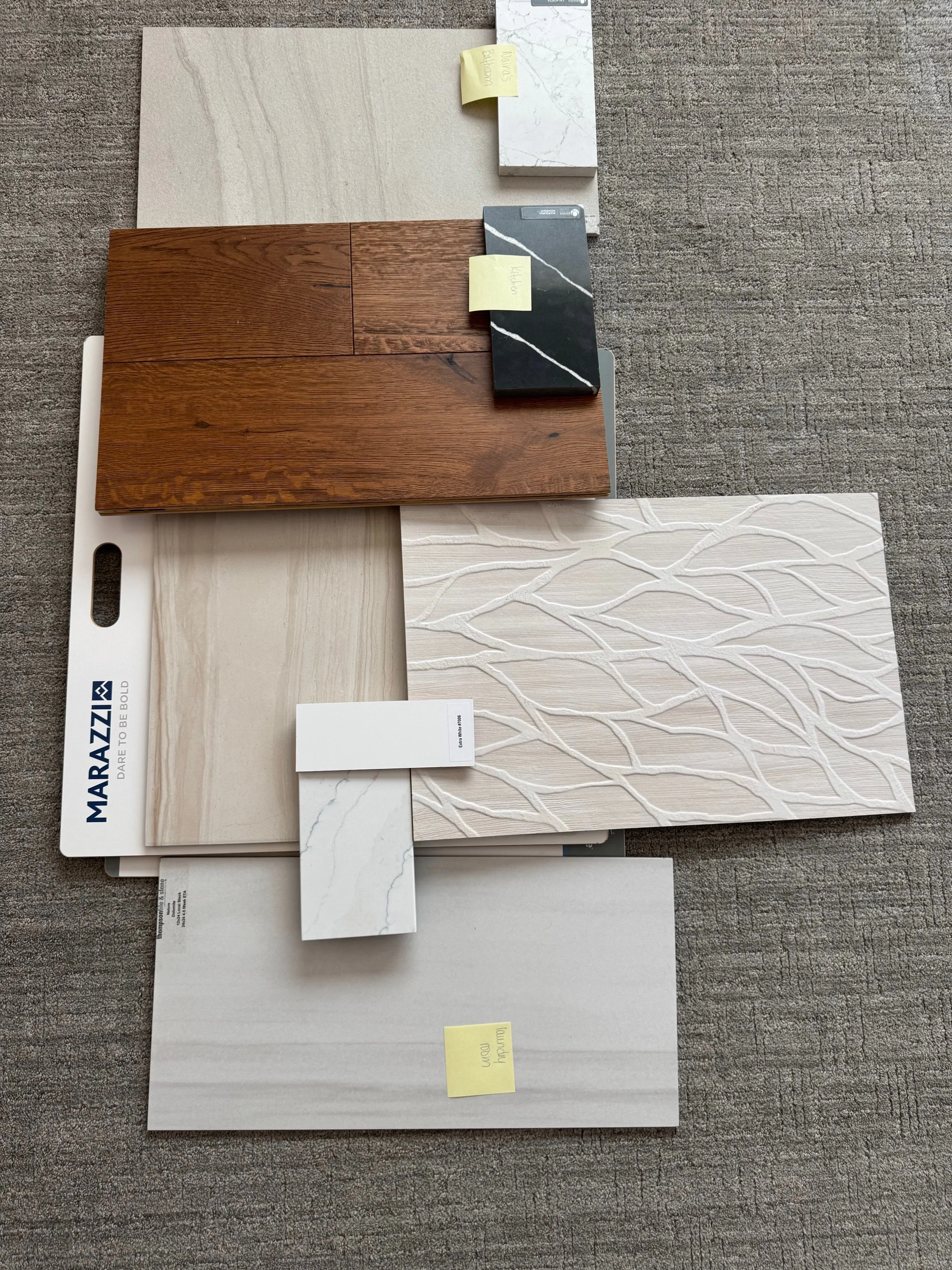 Arrangement of various interior design tiles and samples, including wood, marble, and textured ceramic tiles, on a gray carpet.