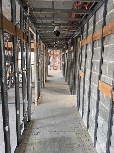 Construction site hallway with metal framing and exposed concrete block walls, unfinished ceiling with pipes and wires, and a concrete floor.