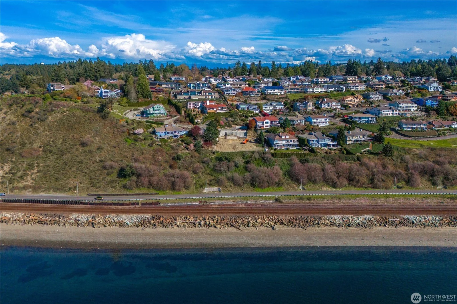 Aerial view of a coastal hillside neighborhood with numerous houses and a train track running along the shoreline with water in the foreground, under a blue sky with clouds.