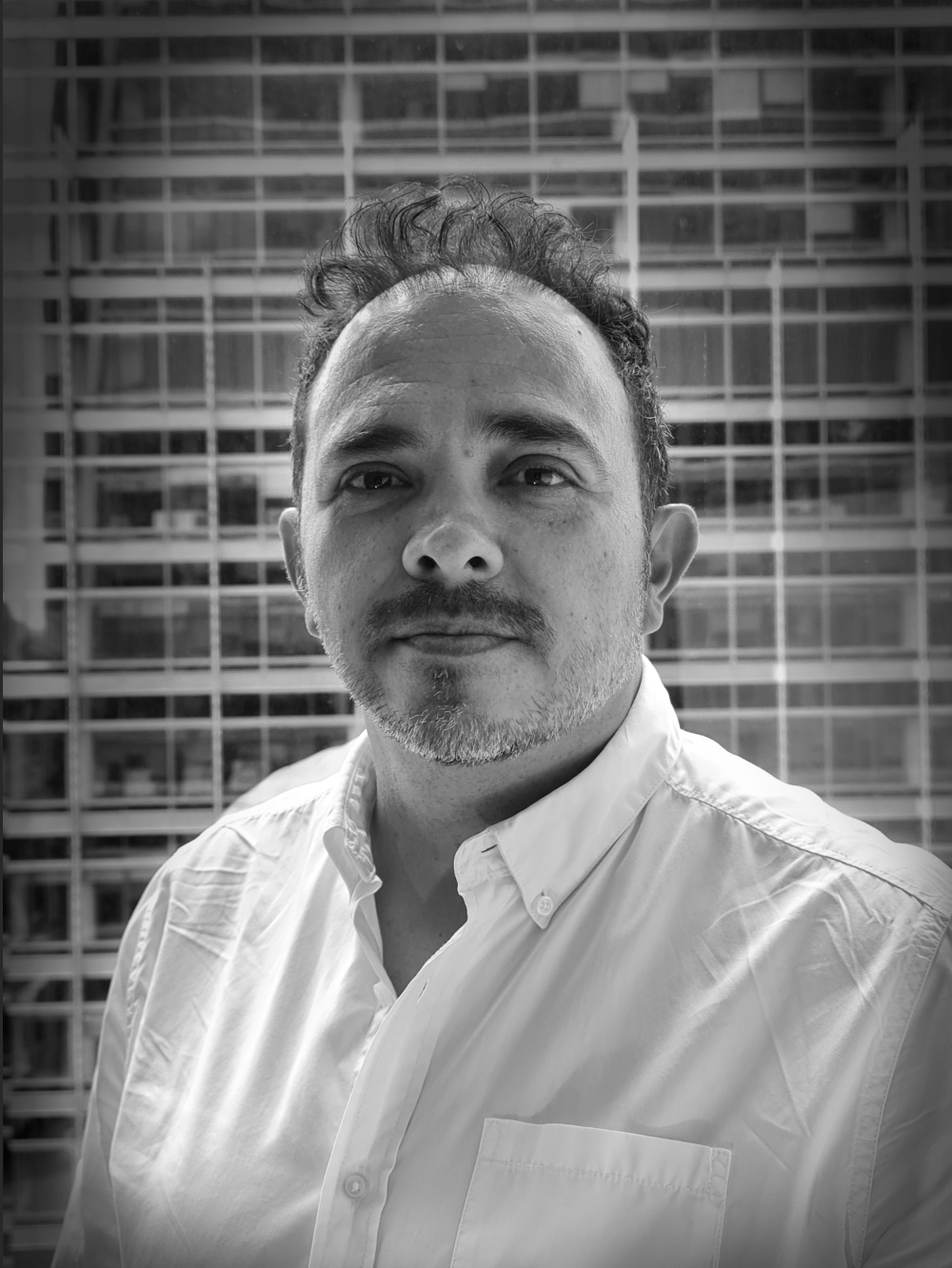A black and white portrait of a man with short curly hair and a beard, wearing a collared shirt, standing in front of a modern glass building.