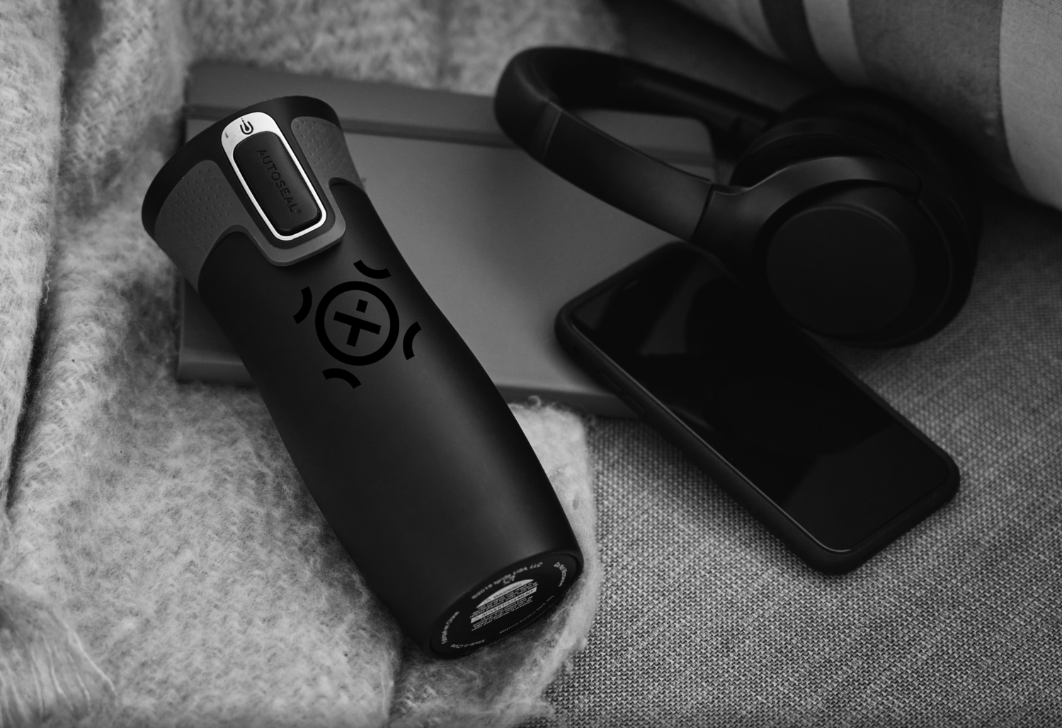 Black noise-canceling headphones, a smartphone, a closed laptop, and a black portable speaker with a logo, all placed on a gray textured surface.