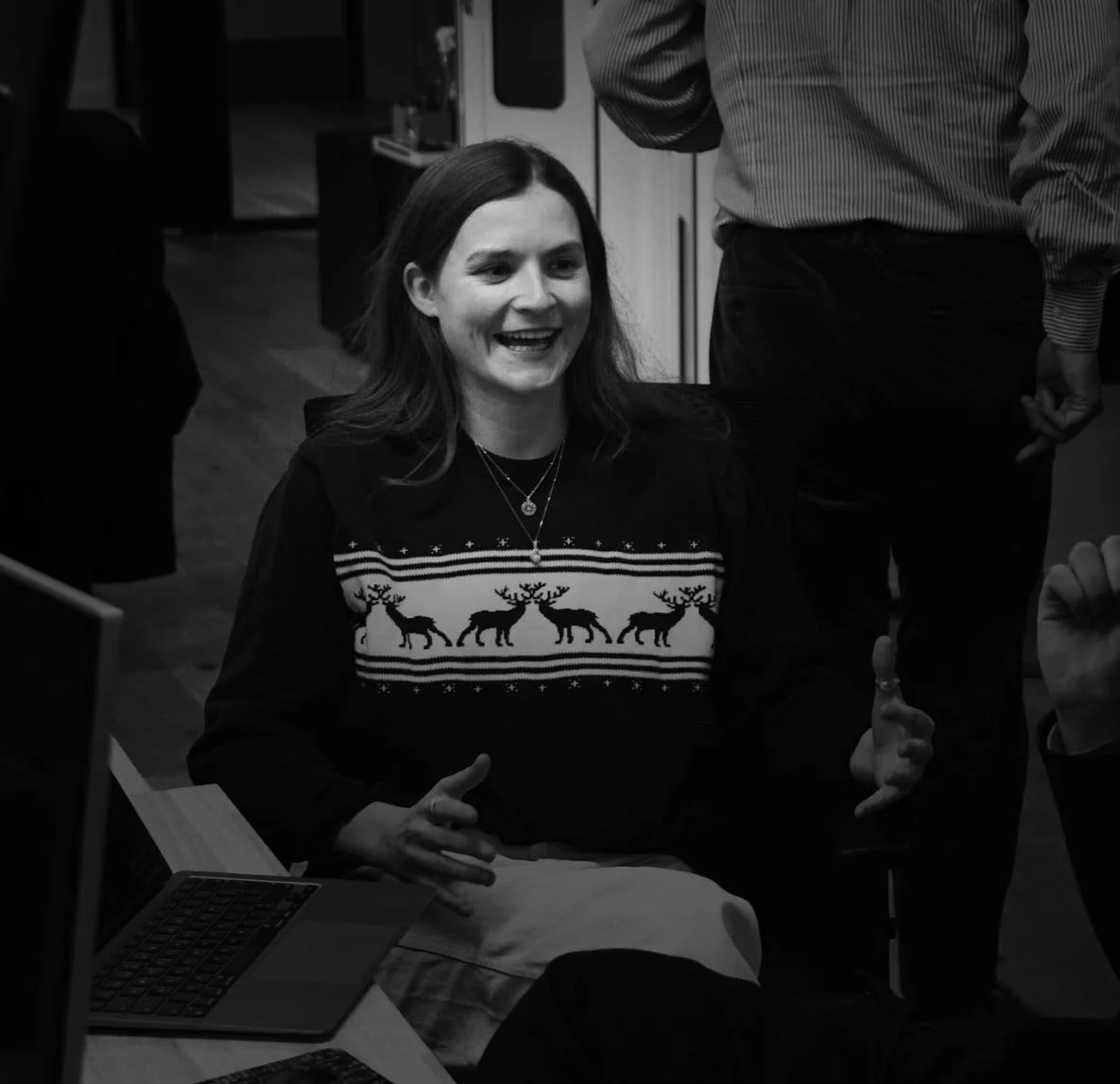 A woman with long hair smiling and talking, wearing an ugly Christmas sweater with reindeer designs, sitting at a desk with a laptop, in a room with other people.