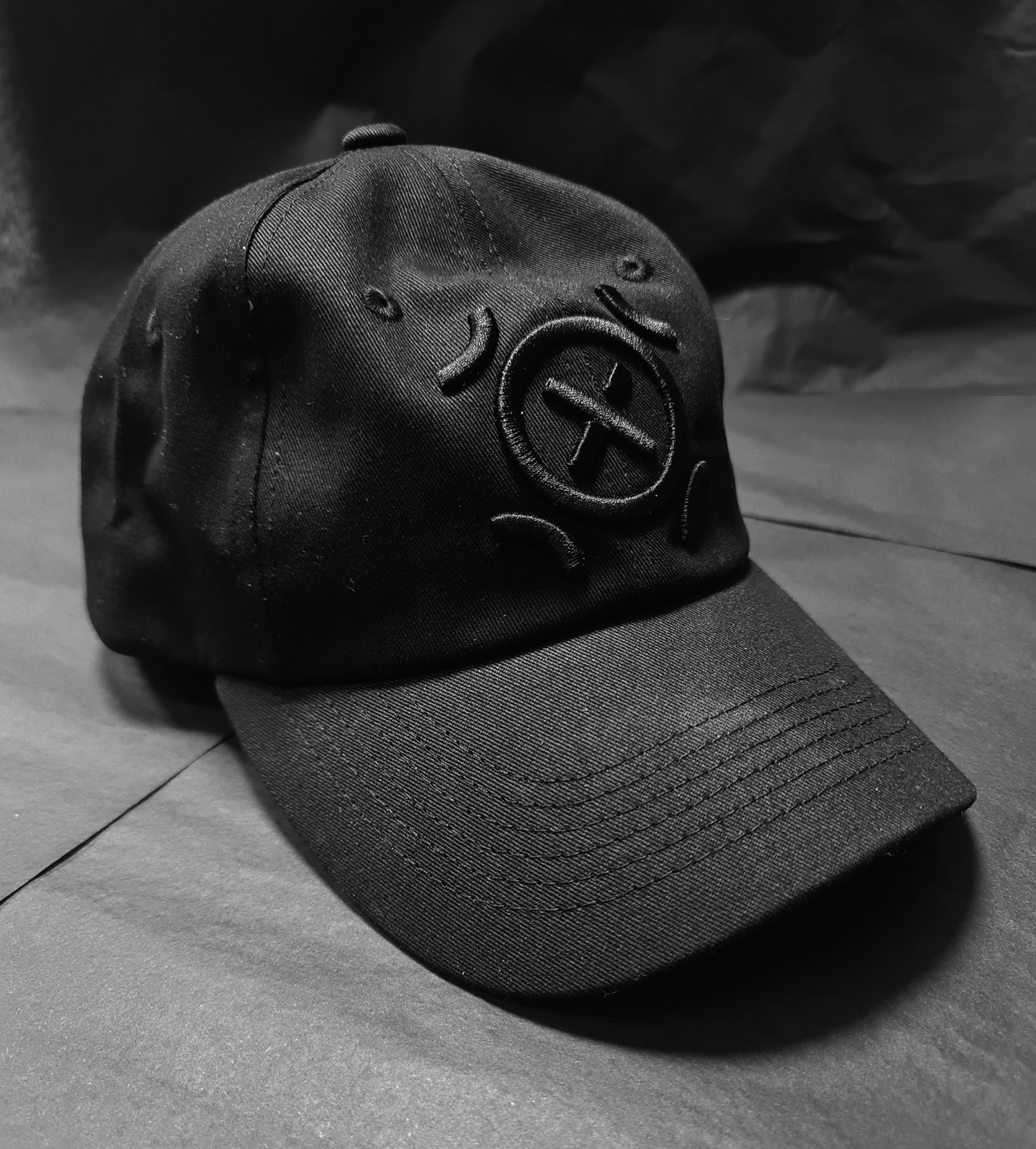 A black baseball cap with embroidered face emojis, including a circle with a crossed-out eye, eyebrows, and a smiling mouth, placed on a flat surface with dark background.