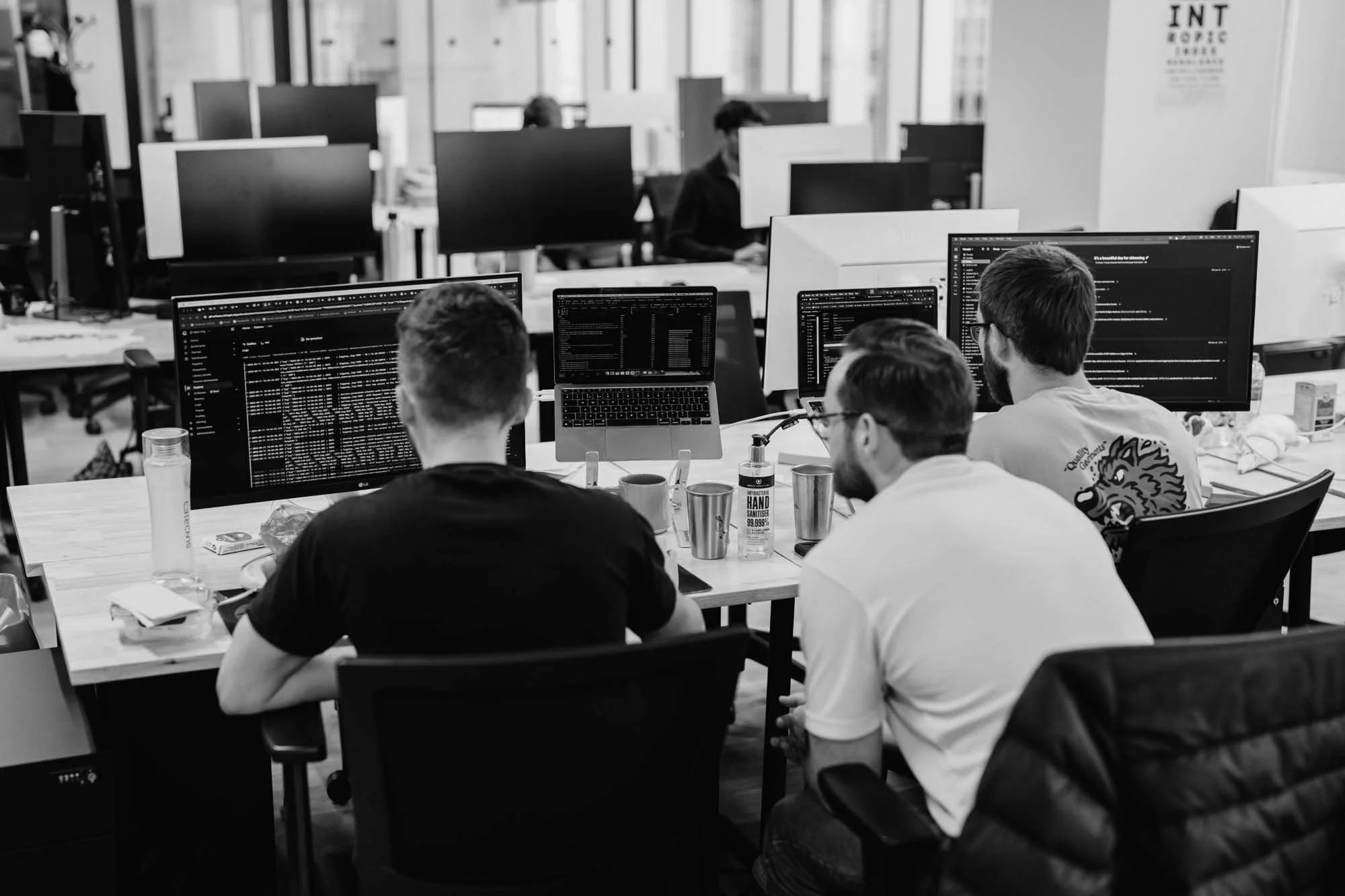 Four men working on computers with multiple monitors in an office environment, focused on code or data on their screens.