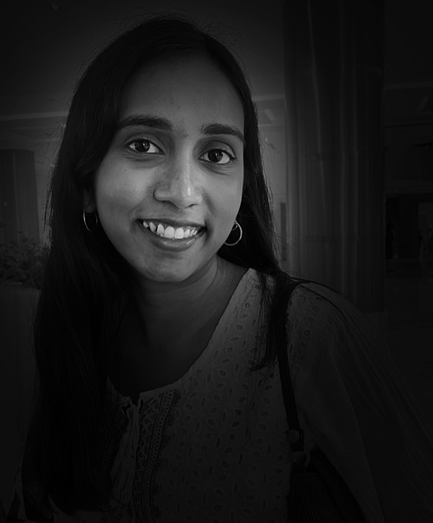 Black and white photo of a smiling woman with long hair, wearing earrings, a patterned top, and carrying a shoulder bag, indoors with curtains and furniture in the background.
